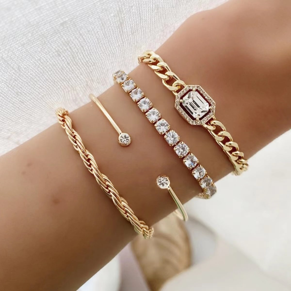 Stackable bracelet set featuring gold designs with twisted rope, bar, tennis, and emerald-cut chain bracelets adorned with cubic zirconia stones.